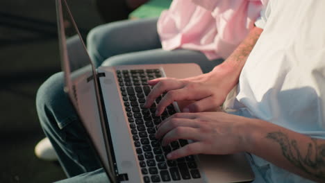 primer plano de manos escribiendo en el teclado de una computadora portátil al aire libre mientras otra persona sostiene un libro en el fondo, con vegetación borrosa y luz solar