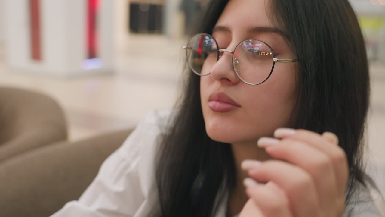 Close up of calm lady resting head on hand while gently adjusting glasses, wearing white shirt with floral tattoo visible, set against softly lit indoor background