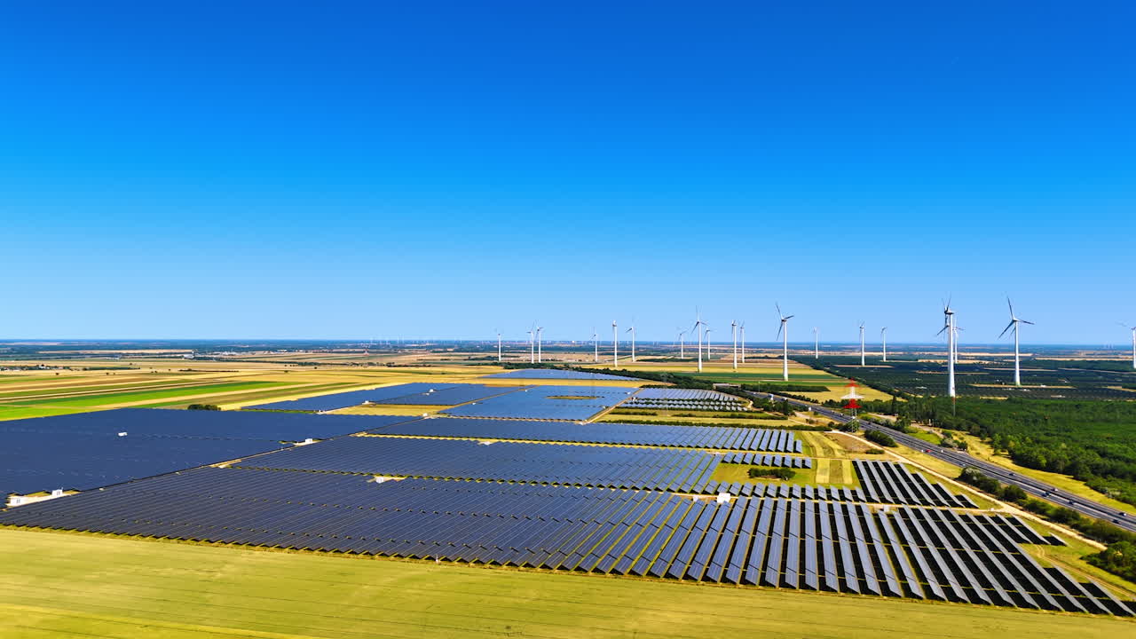 Lots of solar panels installed in the countryside. Wind farms work at backdrop. Aerial view