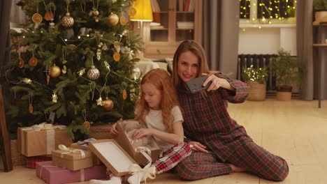 happy young mother and adorable little daughter in pajamas making selfie by decorated christmas tree while sitting on the floor of living room