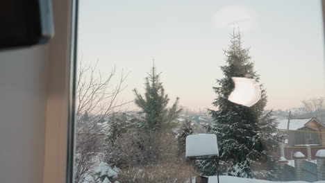 outdoor view of snowy environment featuring pine trees and snow-covered bushes with a well in the compound, rooftops of distant homes, and soft pastel sky during cold winter morning