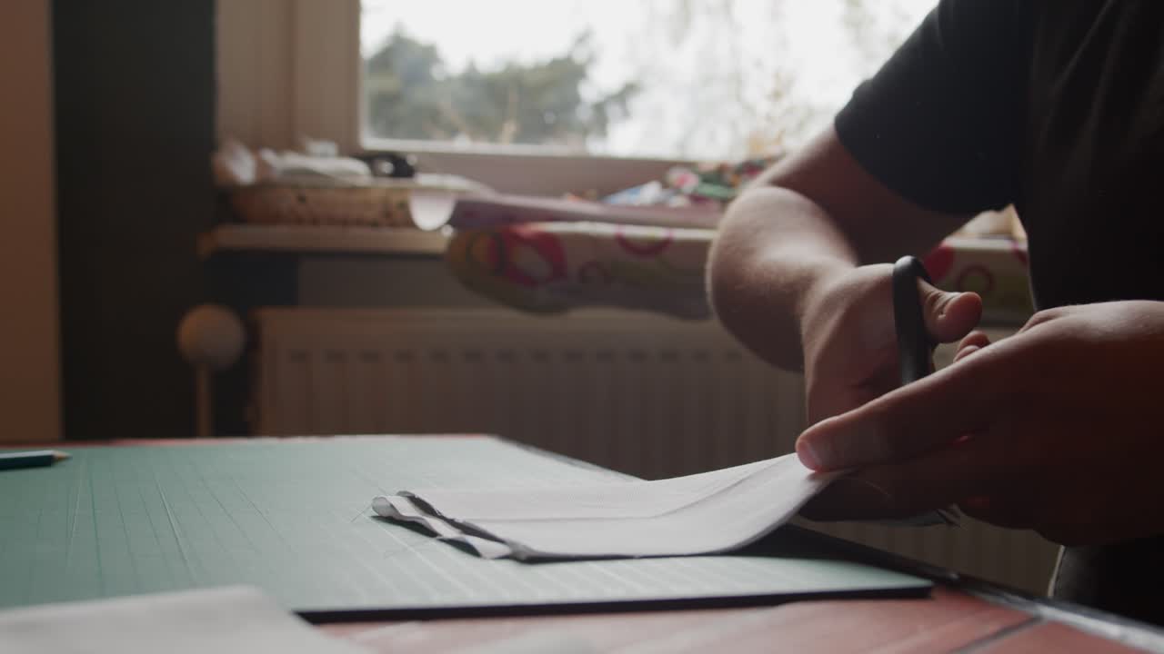 A woman or young person seated cuts fabric in slow motion with a window lighting from behind
