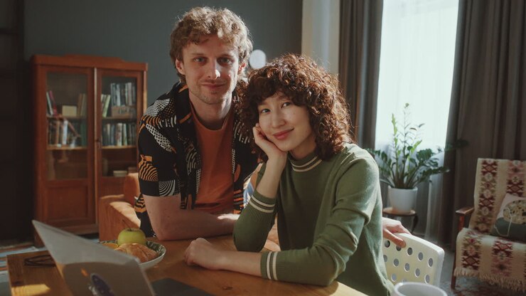 Portrait of Cheerful Couple Sitting at Table with Laptop in Sunlit Living Room