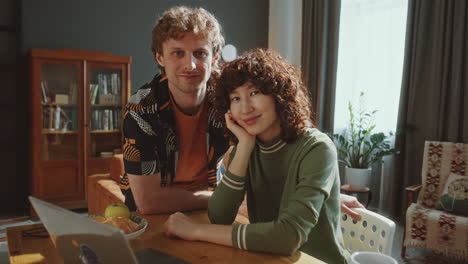 portrait of cheerful couple sitting at table with laptop in sunlit living room