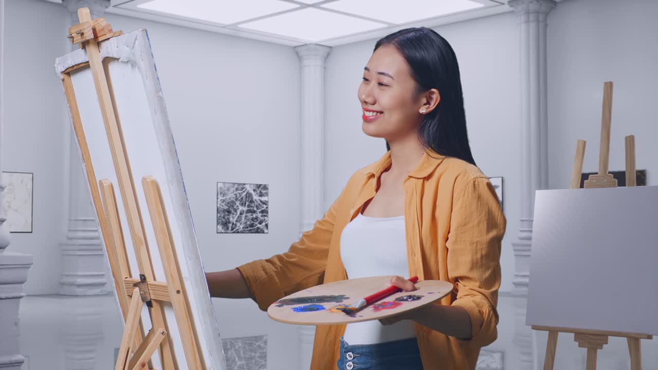 아시아 여성 예술가 는 갤러리 에서 오일 페인트 와 브러쉬 로 캔버스 에 그림 을 그리는 동안 엄지손가락 을 올려 놓고 미소 를 짓고 있다