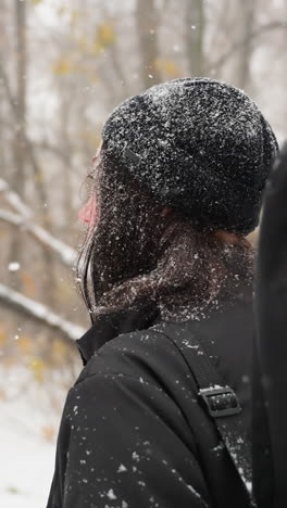 vista lateral de una dama con gorra negra y sudadera con capucha, salpicada de nieve, caminando por un parque cubierto de nieve con una mochila de guitarra, rodeada de árboles desnudos y cargados de helada