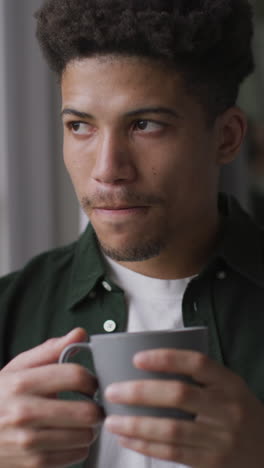 video vertical de un hombre biracial pensativo de pie mirando por la ventana bebiendo café
