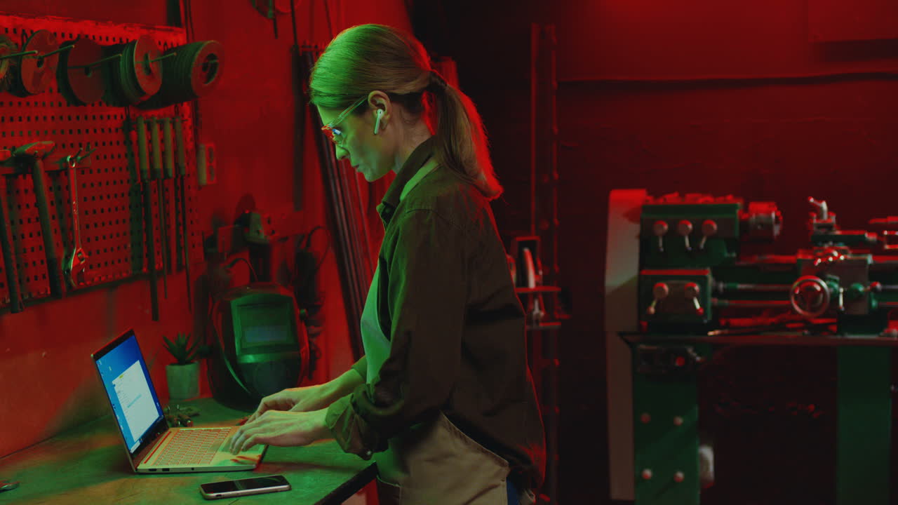 вид сбоку на молодую женщину в фартуке и защитных очках, использующую ноутбук в металлической мастерской