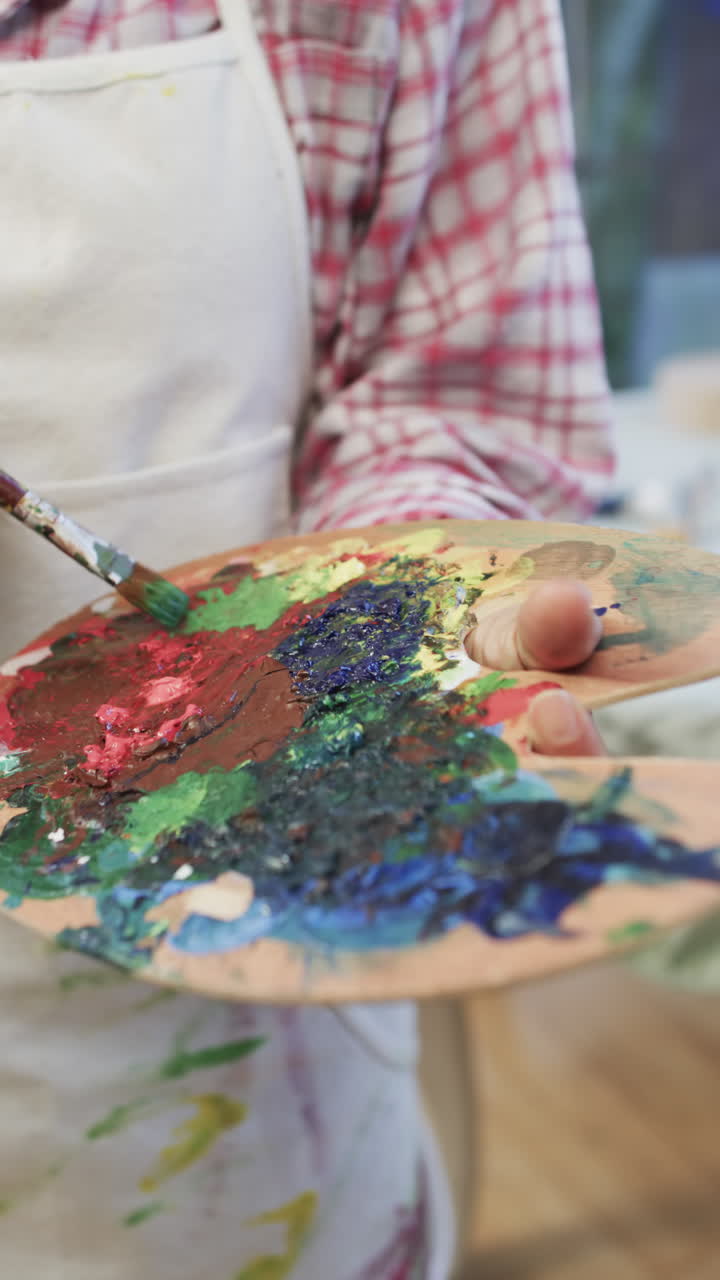 Video verticale della mano di una donna birazziale con grembiule che mescola colori con un pennello in studio, al rallentatore