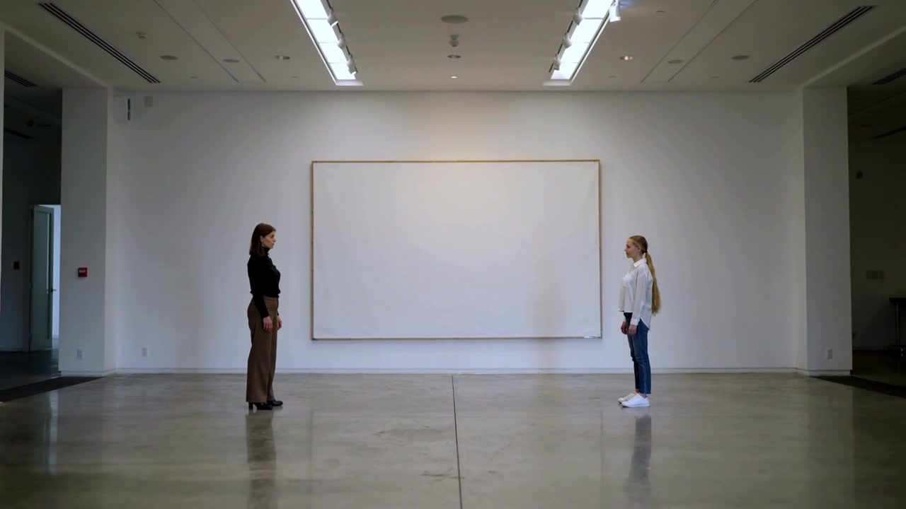 Camera starting, two women in black top white shirt shifting in gallery for exhibit, blank canvas