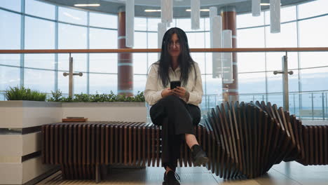 young adult seated cross-legged on decorative wooden bench inside modern building, focused on smartphone with book resting at bench end, surrounded by indoor plants and bright glass wall background