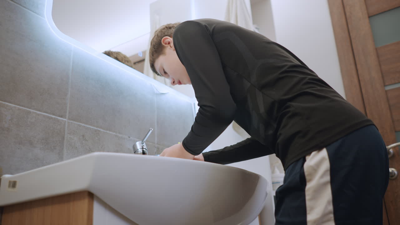 Athletic teen at bathroom sink rinsing face and grabbing towel after activity, bright vanity light, quick refresh routine, sports shorts and short sleeve top, energetic morning vibe
