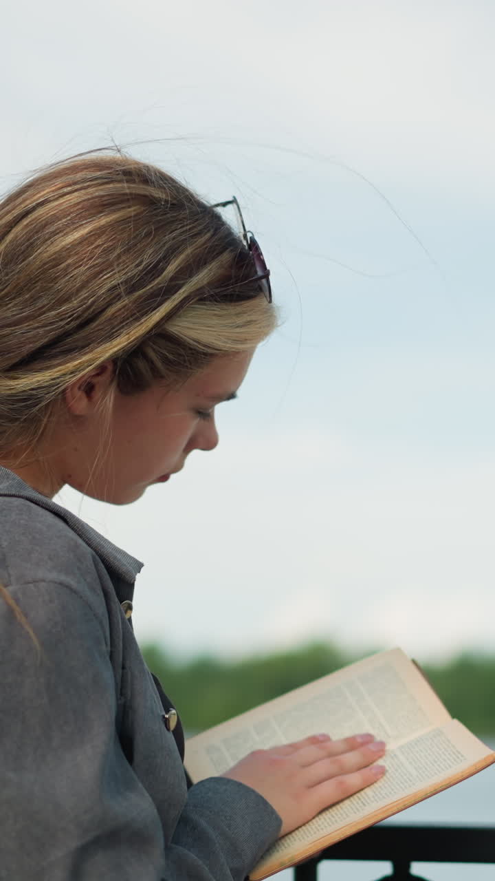 dama mirando a la distancia centrarse de nuevo en su libro mientras volvía a una nueva página de su libro, descansando su mano en una valla de hierro, el fondo presenta vegetación borrosa y un puente sobre el río