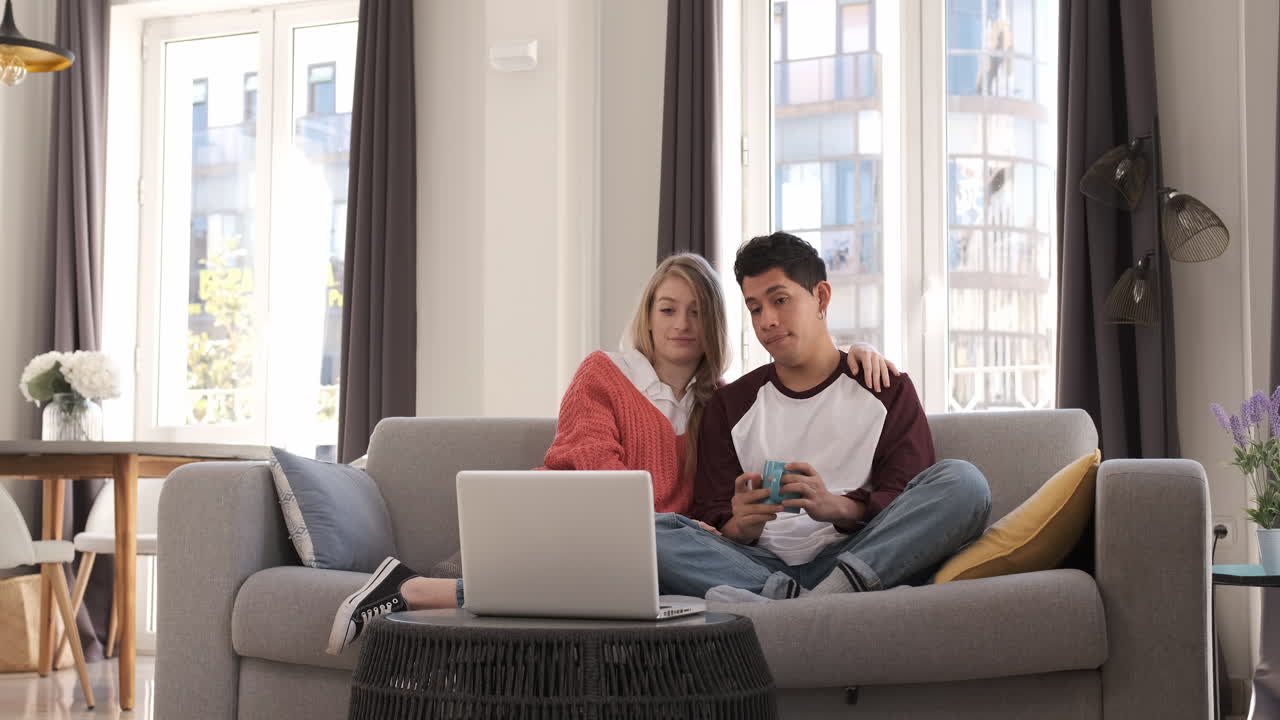 Young couple on a video call with their laptop while sitting on a couch together at home.