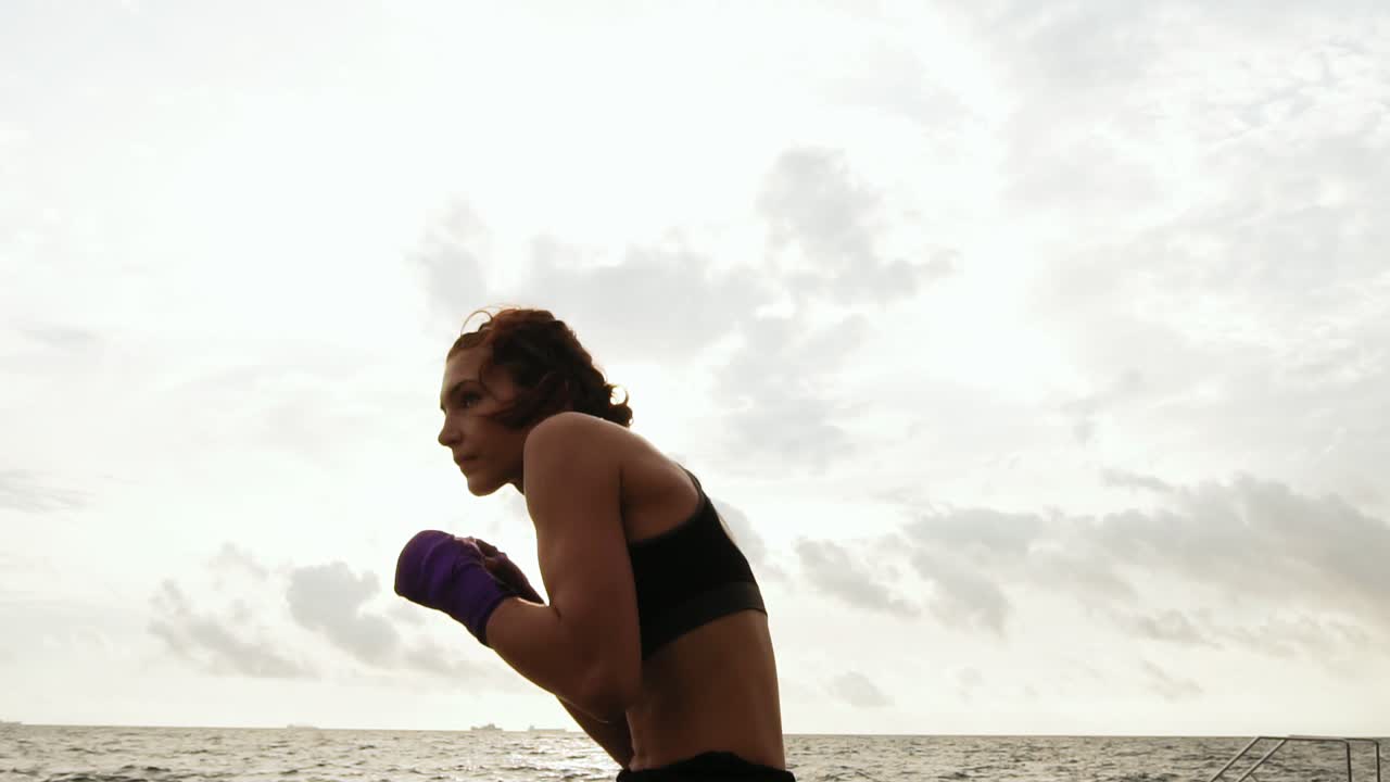 mujer joven atlética shadowboxing por el mar contra el hijo. hermosa mujer boxeadora entrenamiento en la playa por la mañana