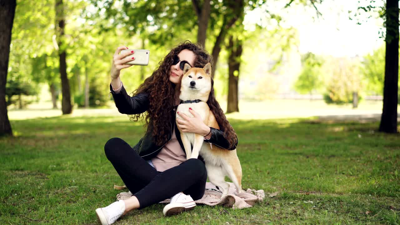 쾌활한 블로거 반려동물 소유자는 스마트폰을 사용하여 개와 셀카를 찍고, 인간과 동물은 공원의 잔디에 앉아 포즈를 취하고, 여성은 개를 만지고 키스하고 있습니다.
