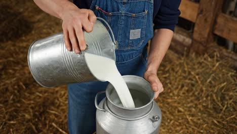 a person pouring fresh milk from a bucket into a traditional milk can in a barn