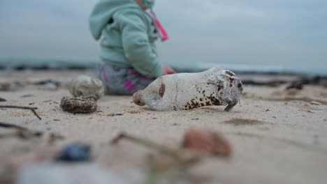 Primer-Plano-De-Una-Botella-De-Plástico-En-Una-Playa-Con-Un-Bebé-Sentado-En-El-Fondo