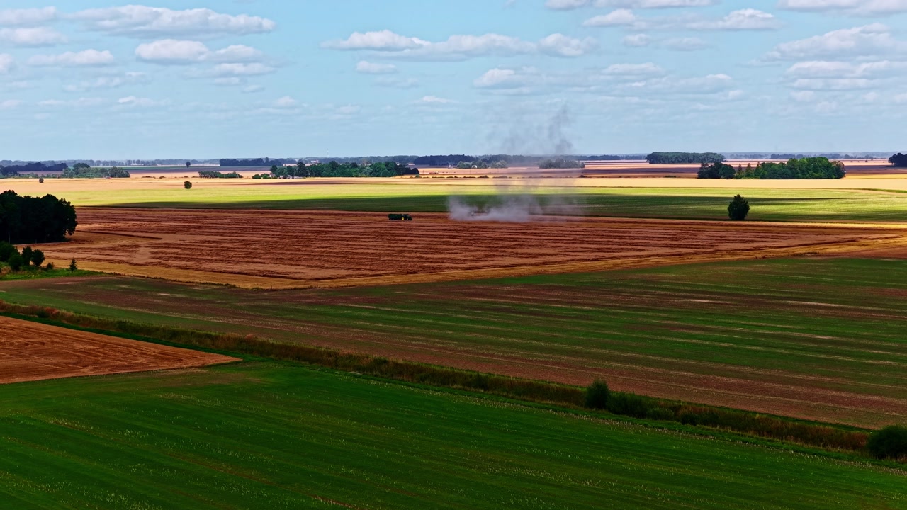 Drone aerial over patchwork farmland with green fields and freshly harvested strips, distant machinery raising light dust under a bright summer sky.