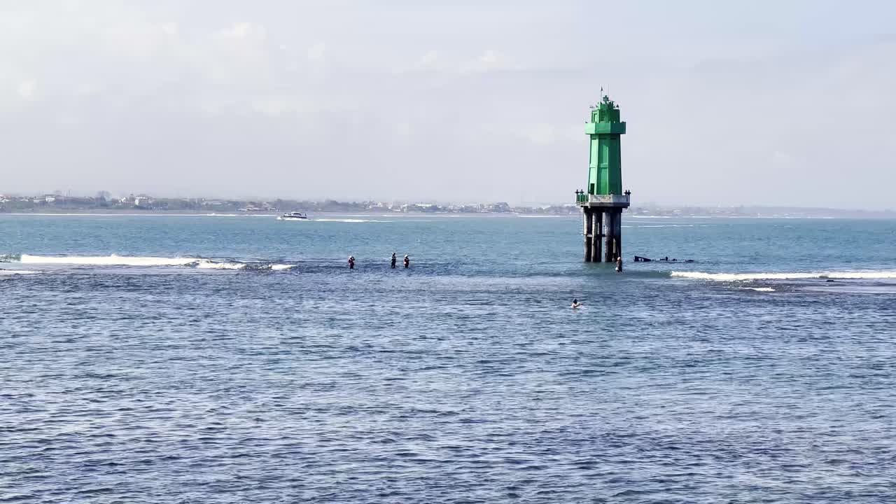 Local fishermen work in the shallow water of the Indonesian sea, with a bright green lighthouse, a symbol of hope, in the background