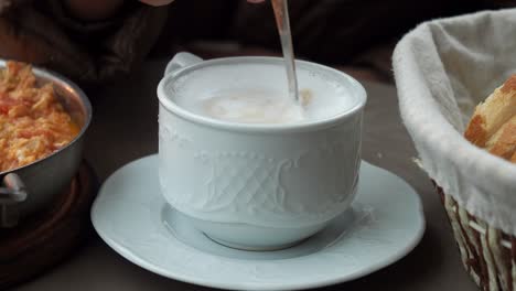 a cup of cappuccino with foam on top, sitting on a white saucer, next to a plate of scrambled eggs and a basket of bread. the image is taken from a table in a cafe.