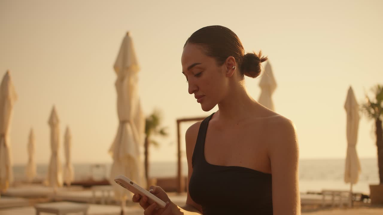 A brunette girl in a black top works on a smartphone and then looks away on a sunny beach in summer. Golden hour on the beach