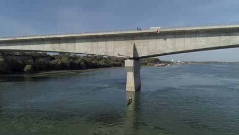 Aerial-HD-shot-showing-a-large-concrete-bridge-over-Danube-in-Belgrade,-Zemun-and-some-fishermen-in-a-small-boat-beneath-on-a-nice-sunny-day