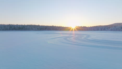 Curving-tyre-tracks-on-Norbotten-Polar-circle-ice-lake-aerial-view-towards-glowing-sunrise-skyline