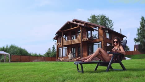 a beautiful slender woman lies on a sun lounger in a swimsuit with a tablet on the background of a large country house in sunglasses and makes online purchases