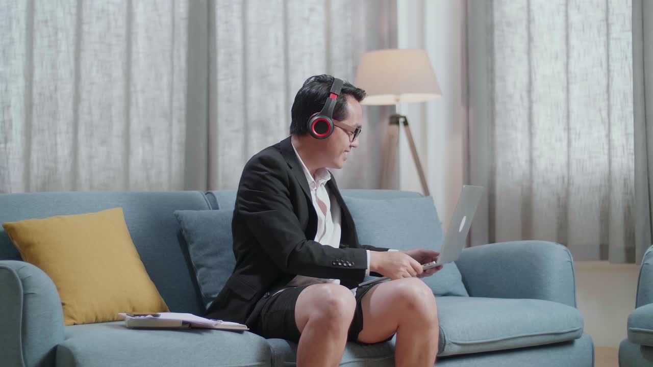 재과 쇼트를 입은 아시아 사업가 가 헤드폰으로 음악을 듣고 집에서 노트북으로 일한 후 춤을 춘다