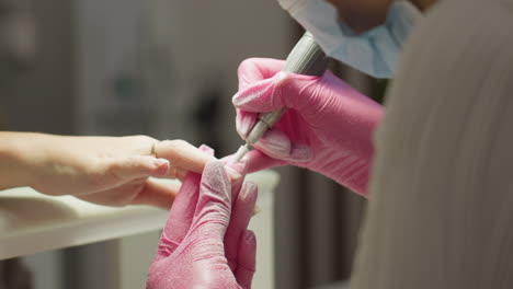 close-up of nail technician in pink gloves filing client nail while wearing protective mask and client wears gold ring and rests hand on surface with soft background blur and flying nail dust