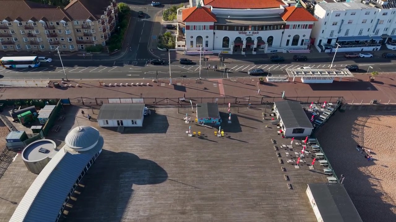 Volo in avanti di un drone attraversa il ponte in legno del molo di Hastings verso la strada, passando il Padiglione Occidentale restaurato con le sue capsule bianche curve, parte della riprogettazione pluripremiata dello storico molo