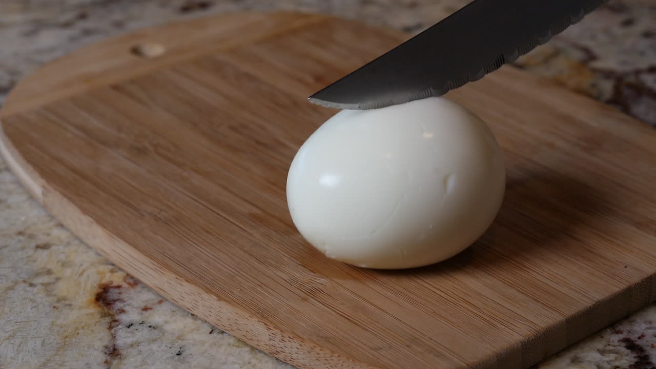 Cutting a hard boiled egg in half with a knife and exposing the yellow egg yolk on top of a wooden cutting board