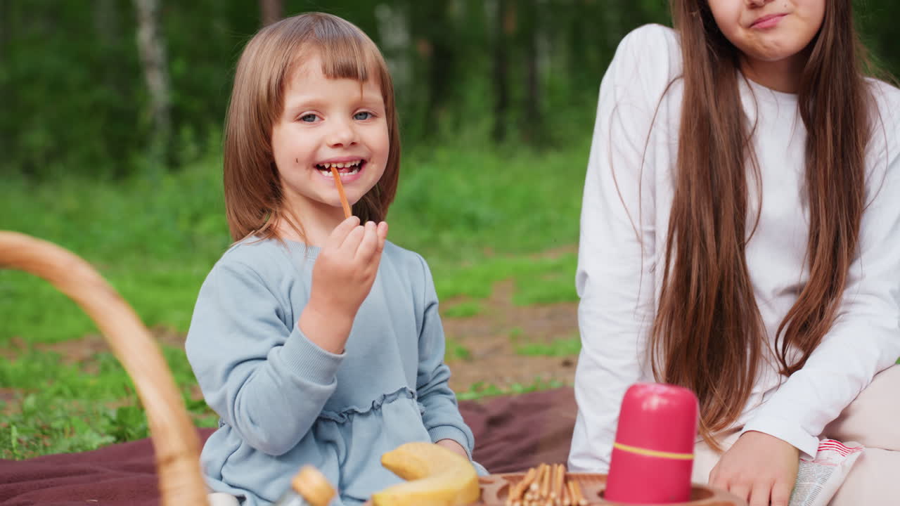 Niña pequeña sentada en una esterilla de pícnic junto a su hermana, comiendo palitos de pan y sonriendo alegremente, mientras su hermana le limpia los restos de los labios, rodeadas de una cesta con aperitivos de pícnic y fondo verde.