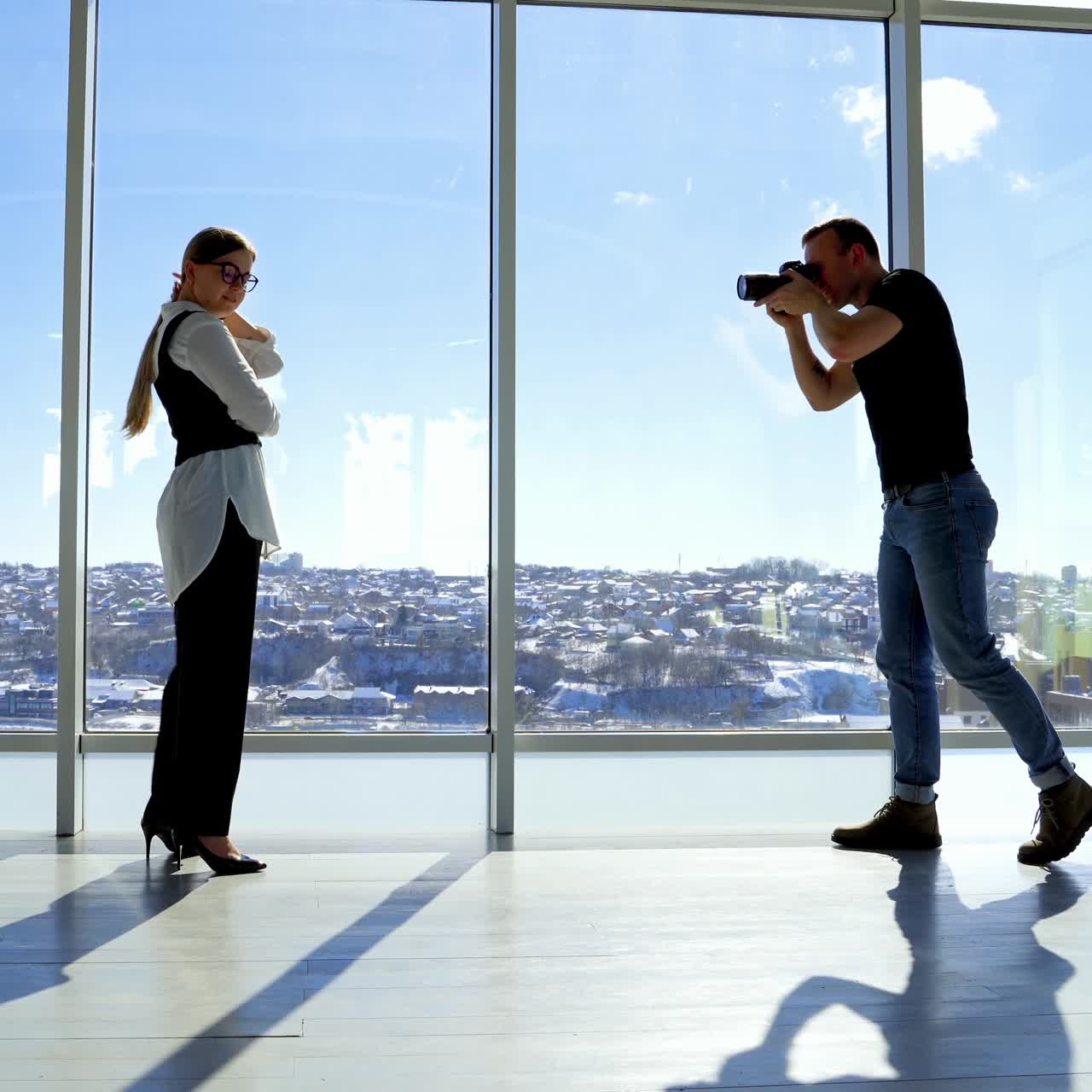 Junge Geschäftsfrau posiert im Büro vor der Kamera. Fotograf macht Fotos von einer Frau in schwarz-weißem Kostüm vor einem Panoramafenster mit Stadtlandschaft.