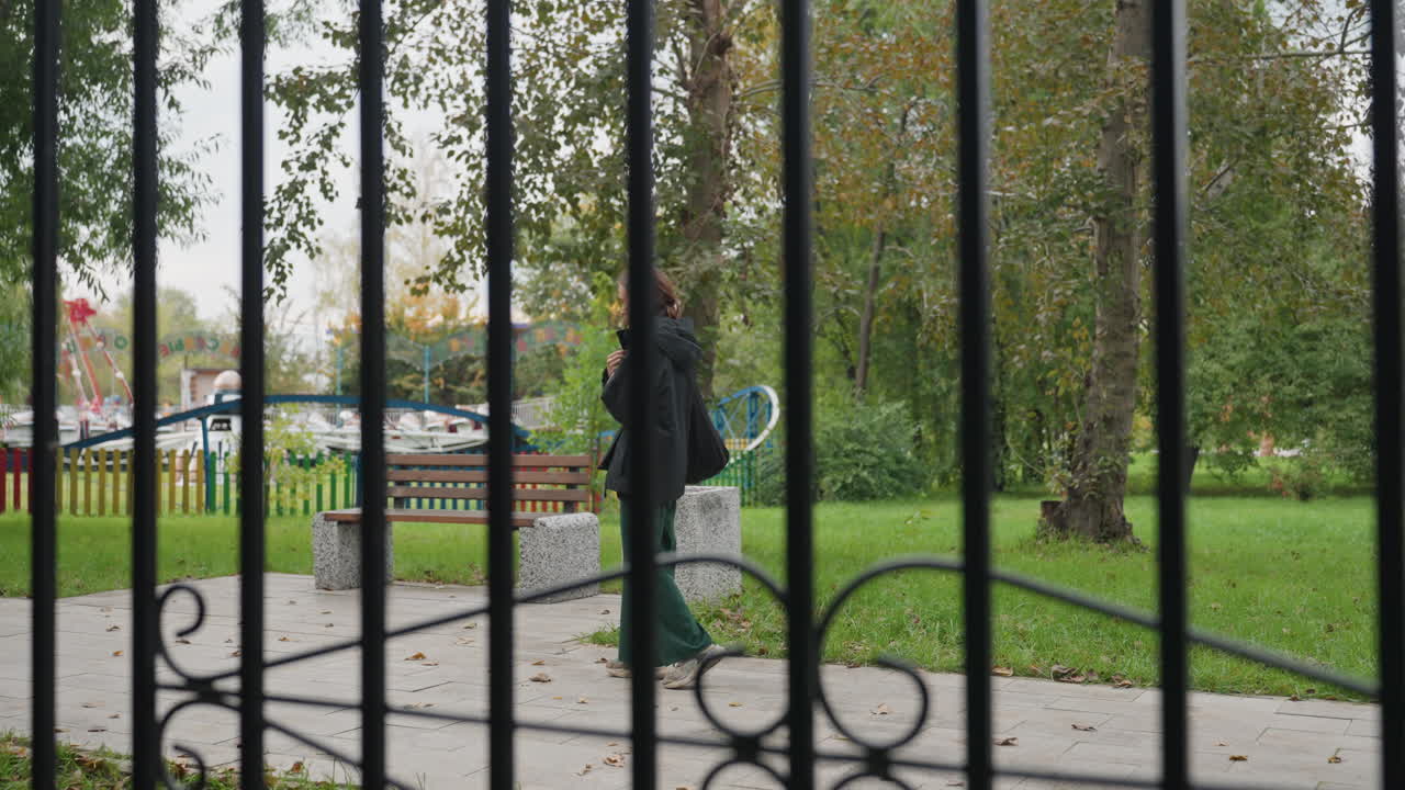 Donna caucasica che cammina dietro una recinzione in un parco autunnale, telefono all'orecchio, atmosfera da pendolare solitaria, indossa un cappotto scuro, supera una panchina e alberi di betulla, le sbarre di ferro in primo piano creano una barriera incorniciata, cielo nuvoloso, silenzio