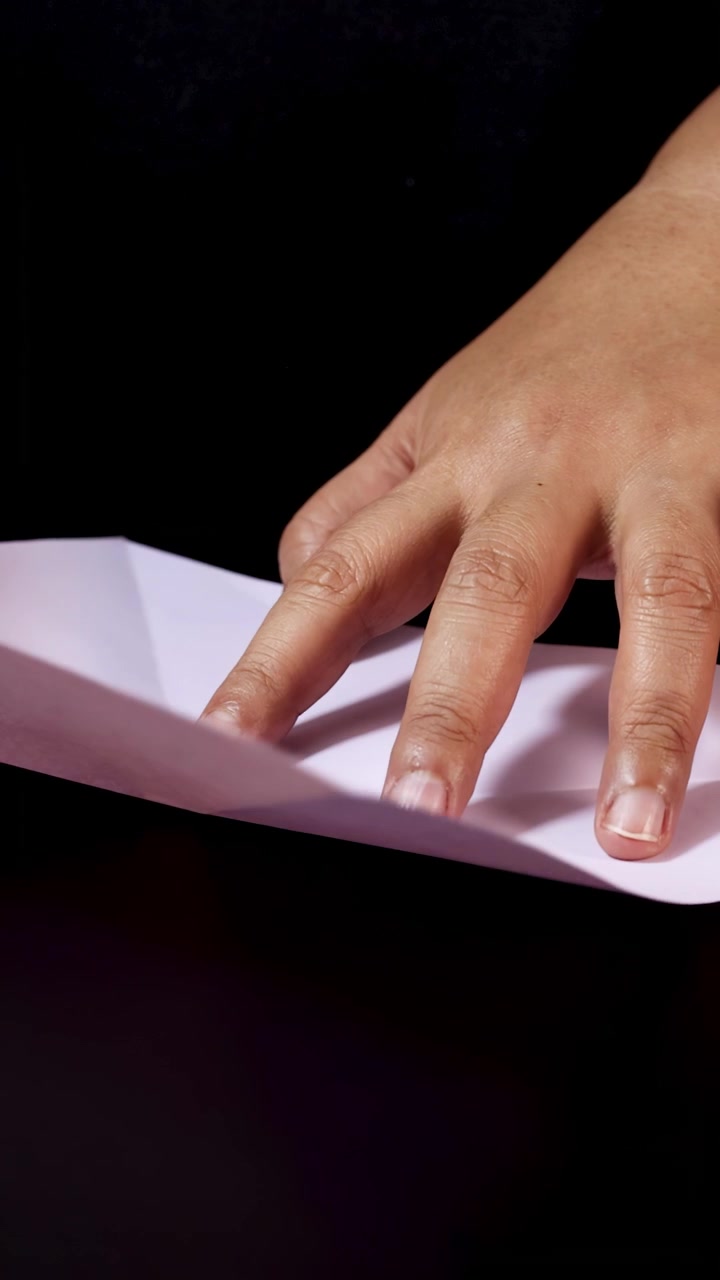 Adult hands carefully fold pastel paper on dark surface, demonstrating step-by-step origami preparation