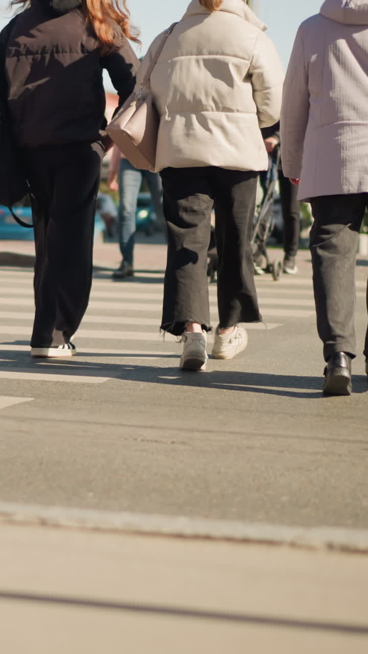 Gruppo che attraversa un passaggio pedonale urbano con piumini e borsette, vista posteriore di amici in scarpe da ginnastica che camminano nella luce autunnale, scena spontanea di pendolarismo con traffico sfocato e vivace atmosfera del centro