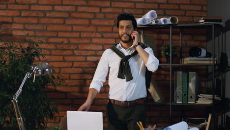 good looking arabian businessman speaking on the phone while standing in the office, then sits at the table with laptop computer