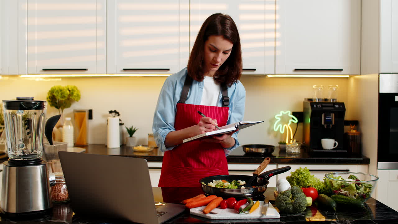 Giovane donna che impara a cucinare sano dal laptop, prendendo appunti, nella moderna cucina di casa.