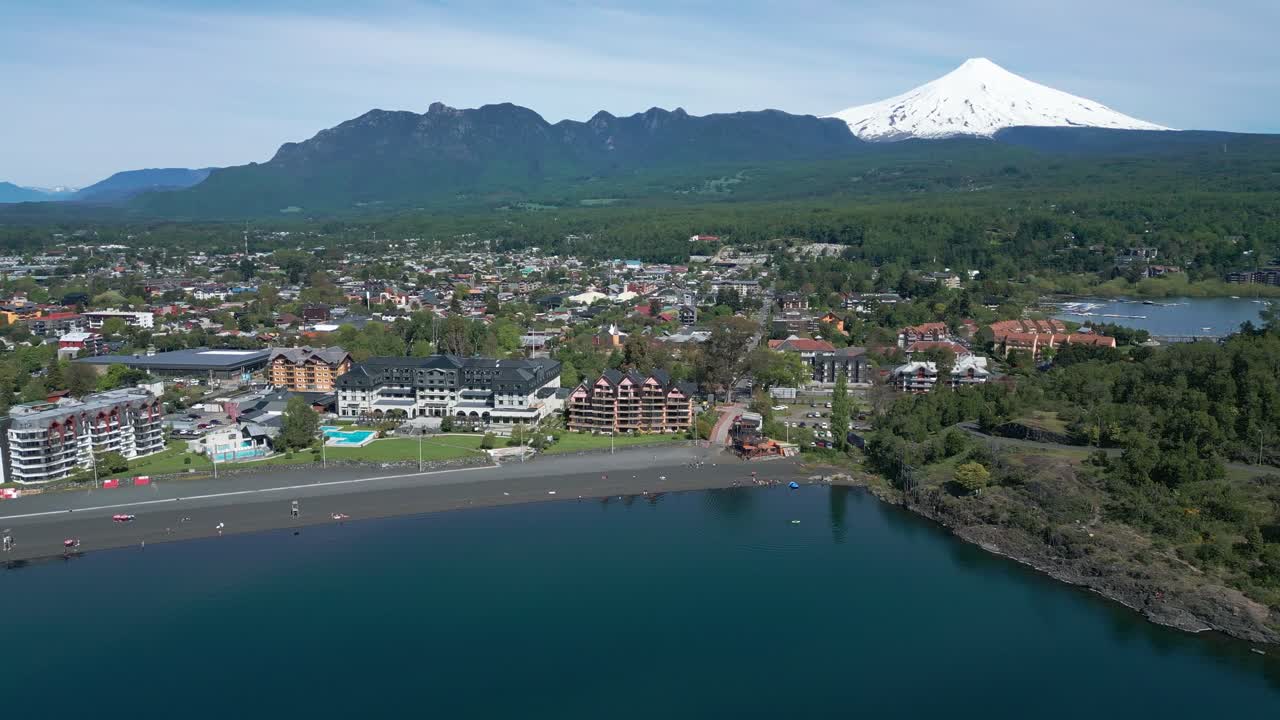 Panoramic drone shot over beach, modern hotels, and black sand, revealing Pucon town and the Villarrica Volcano in the background