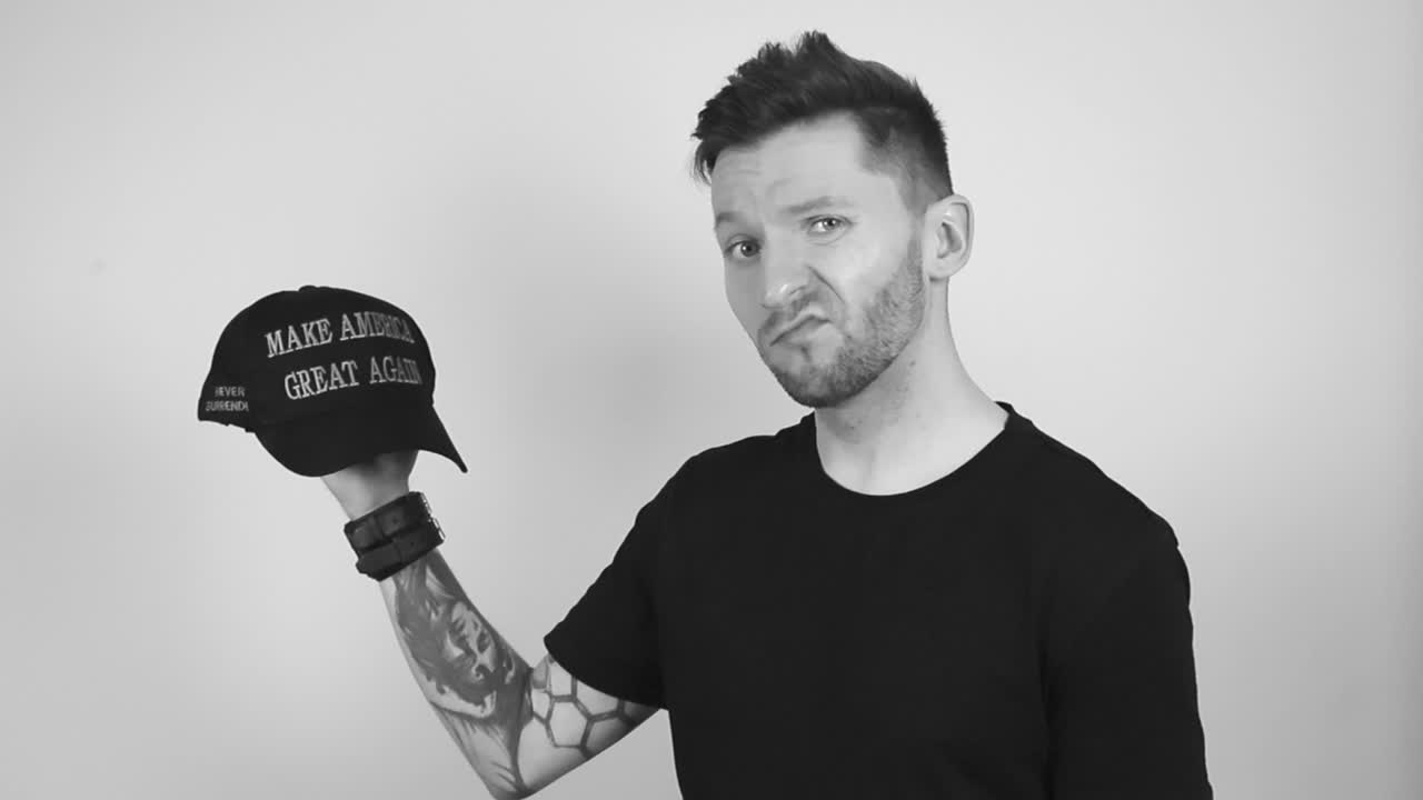 Grayscale footage of a tattooed short haired and bearded man lifting up a black MAGA or hat with the writing Make America Great Again slowly and beings shaking his head with not happy emotions.