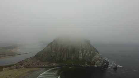 aerial reveal of morro rock in california from the clouds. the seastack towers over the beach and ocean below, contrasted by the fog obscuring its true size. waves calmly cascade onto the beach.