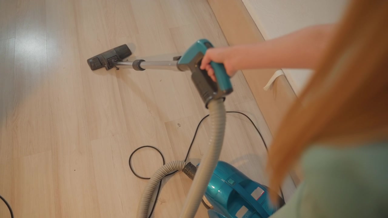 Mulher branca aspirando piso de madeira com aspirador de pó tipo canister azul e fio visível, close-up em perspectiva por cima do ombro mostrando rodapé e remoção de poeira, luz calma da manhã, proprietária realizando rotina