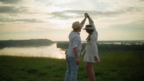 a man spins his wife around on a grassy hill beside a peaceful lake at sunset. the man, dressed in a white shirt, hat, and jeans, as the woman in a white dress and black hat hugs him warmly