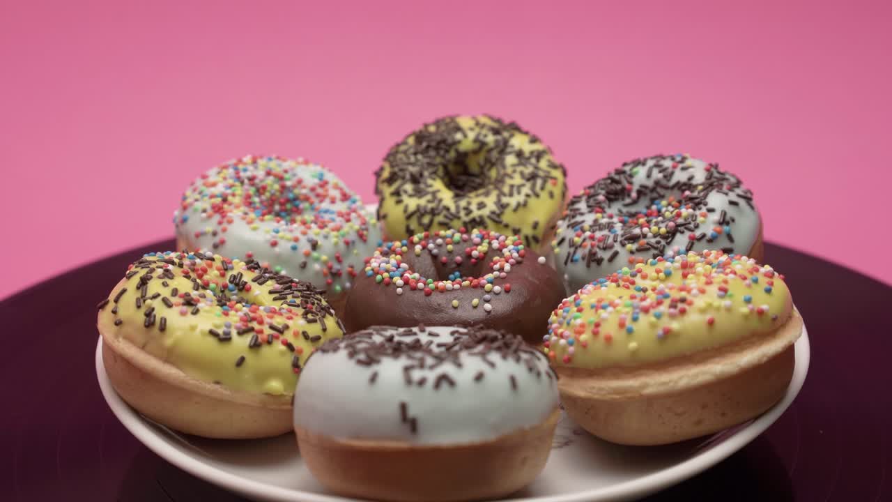 A bunch of American donuts with black, yellow and blue chocolate and choco and multicolored crumb toppings rotates on a pink background