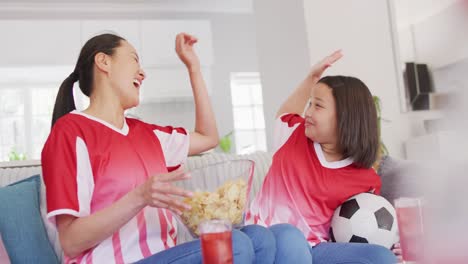 Video-Einer-Glücklichen-Asiatischen-Mutter-Und-Tochter,-Die-Chips-Isst-Und-Sich-Ein-Spiel-Im-Fernsehen-Ansieht