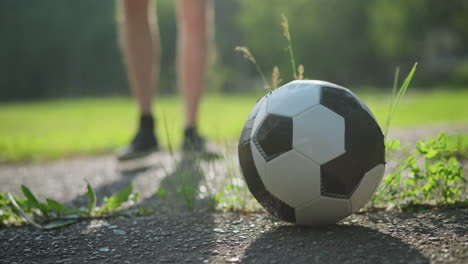 a blurred view of someone wearing brown shorts walking towards a soccer ball placed on a paved surface, with a lush grassy field in the background