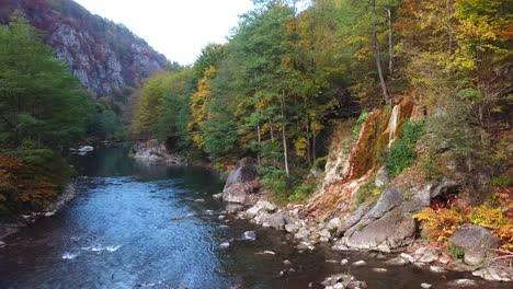 drone shot of a small waterfall flowing down a rocky cliff, surrounded by autumn trees