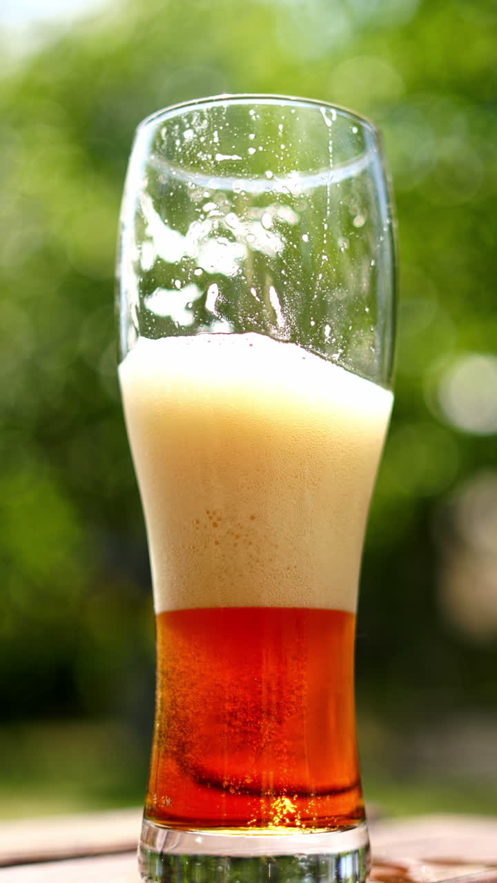 Half glass of cold beer with white foam outdoors. Close-up dark beer with bubbles on blur green background in summer. Vertical video